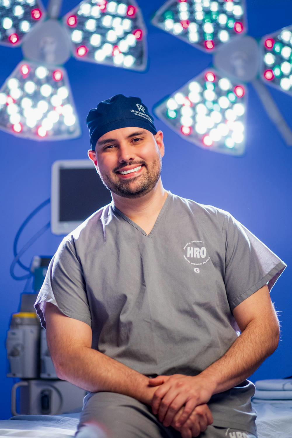 Dr. Ricardo Penteado sorrindo, sentado no centro cirúrgico, com os focos de luz acesos ao fundo